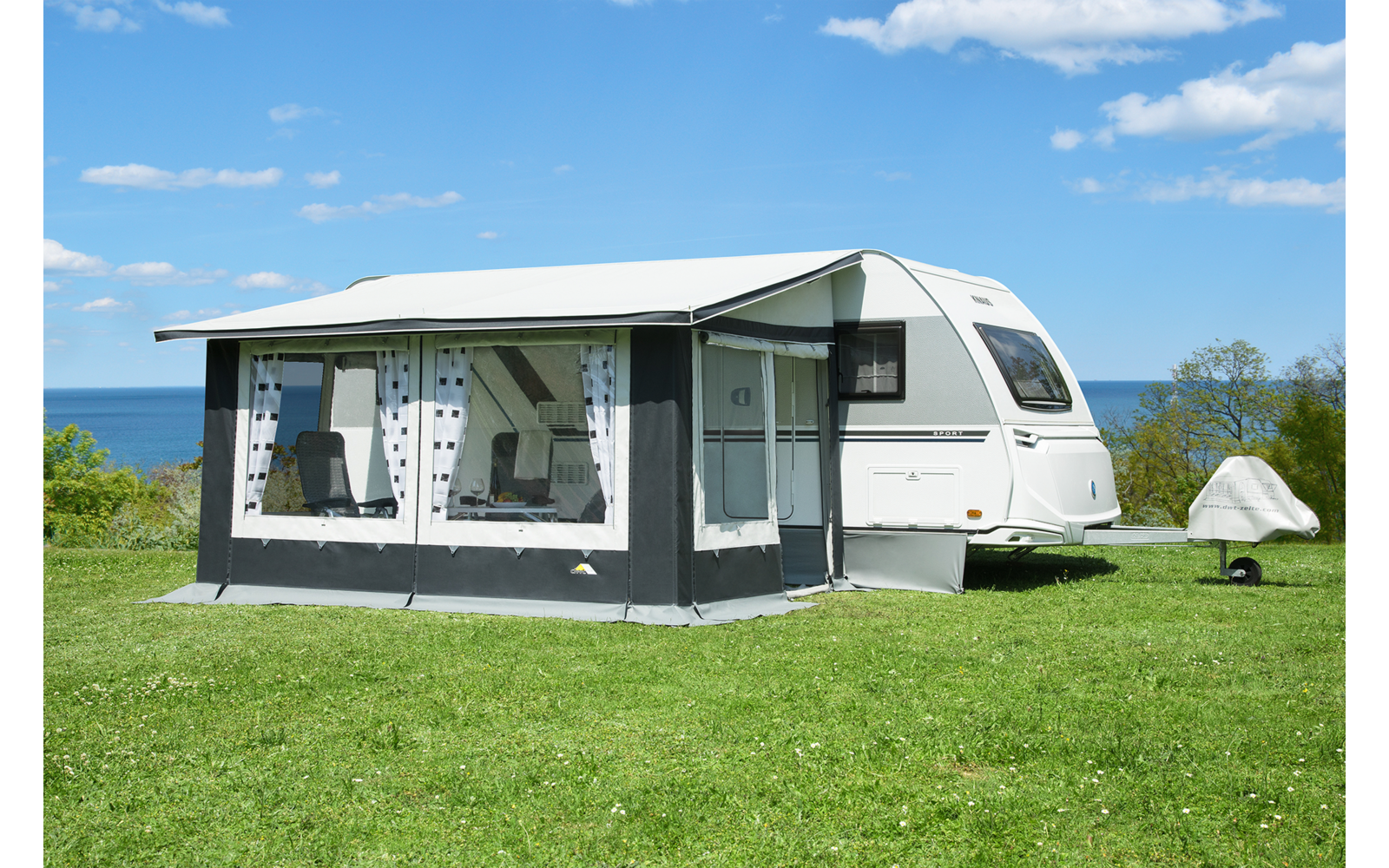Weißer Wohnwagen mit angebautem Vorzelt auf grüner Wiese, im Hintergrund Meer und blauer Himmel mit Wolken