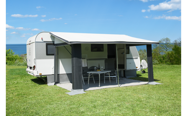 Weißer Wohnwagen auf grüner Wiese, ausgeklappte Markise, darunter ein Tisch mit zwei Stühlen, im Hintergrund Meer und blauer Himmel mit Wolken