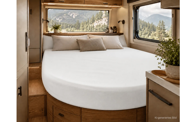 Interieur van een camper met een afgerond bed met wit hoeslaken, drie kussens, houten kasten, ramen met uitzicht op bosrijke bergen en planten op planken