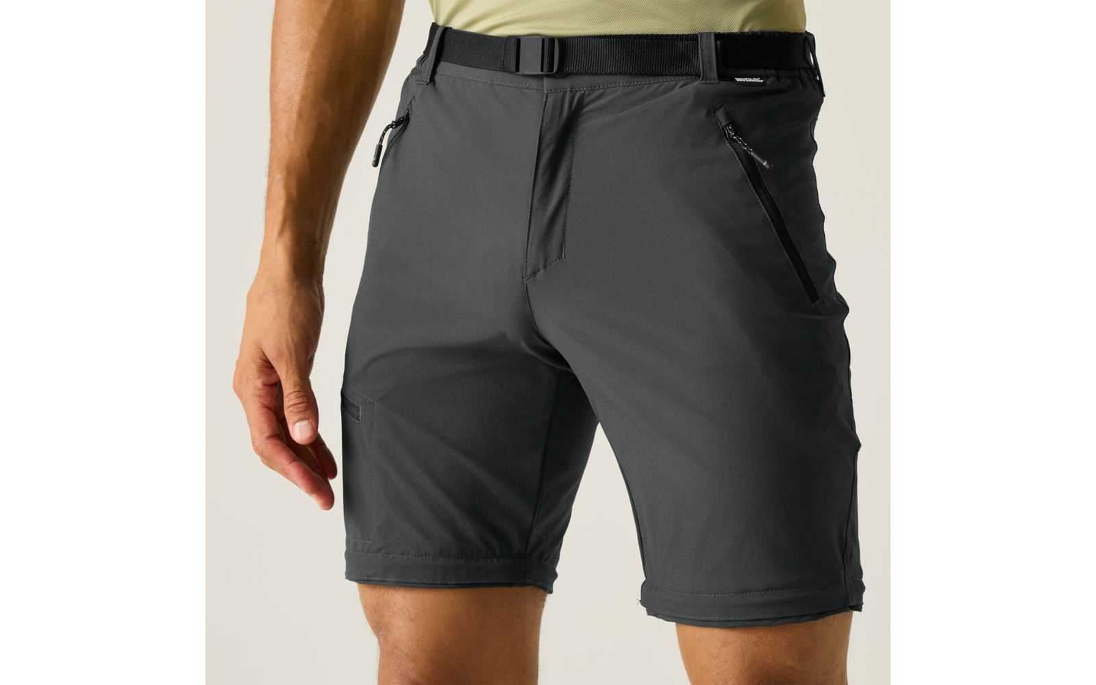 Unterer Rumpf einer Person mit dunkelgrauen Shorts mit Reißverschlusstaschen und Gürtel, hellgrünes T‑Shirt, Hände und Oberschenkel sichtbar, beiger Hintergrund