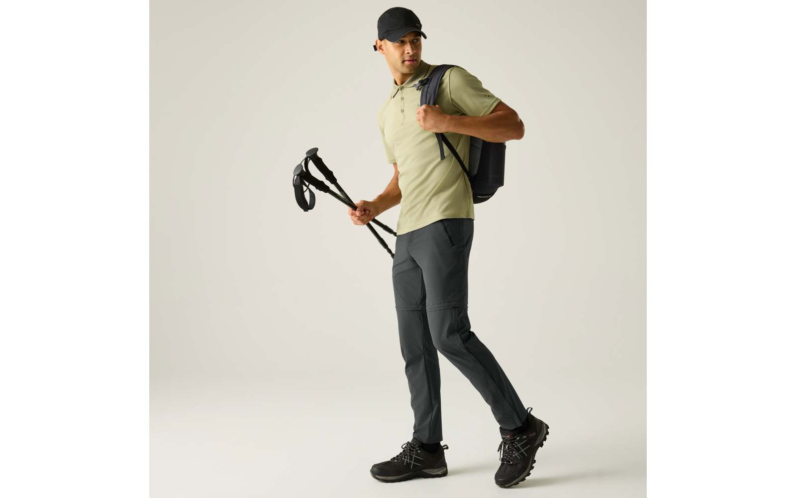 Mann mit schwarzer Kappe, hellgrünem Poloshirt und dunkler Hose, trägt Rucksack und Wanderstöcke, geht und blickt zur Seite