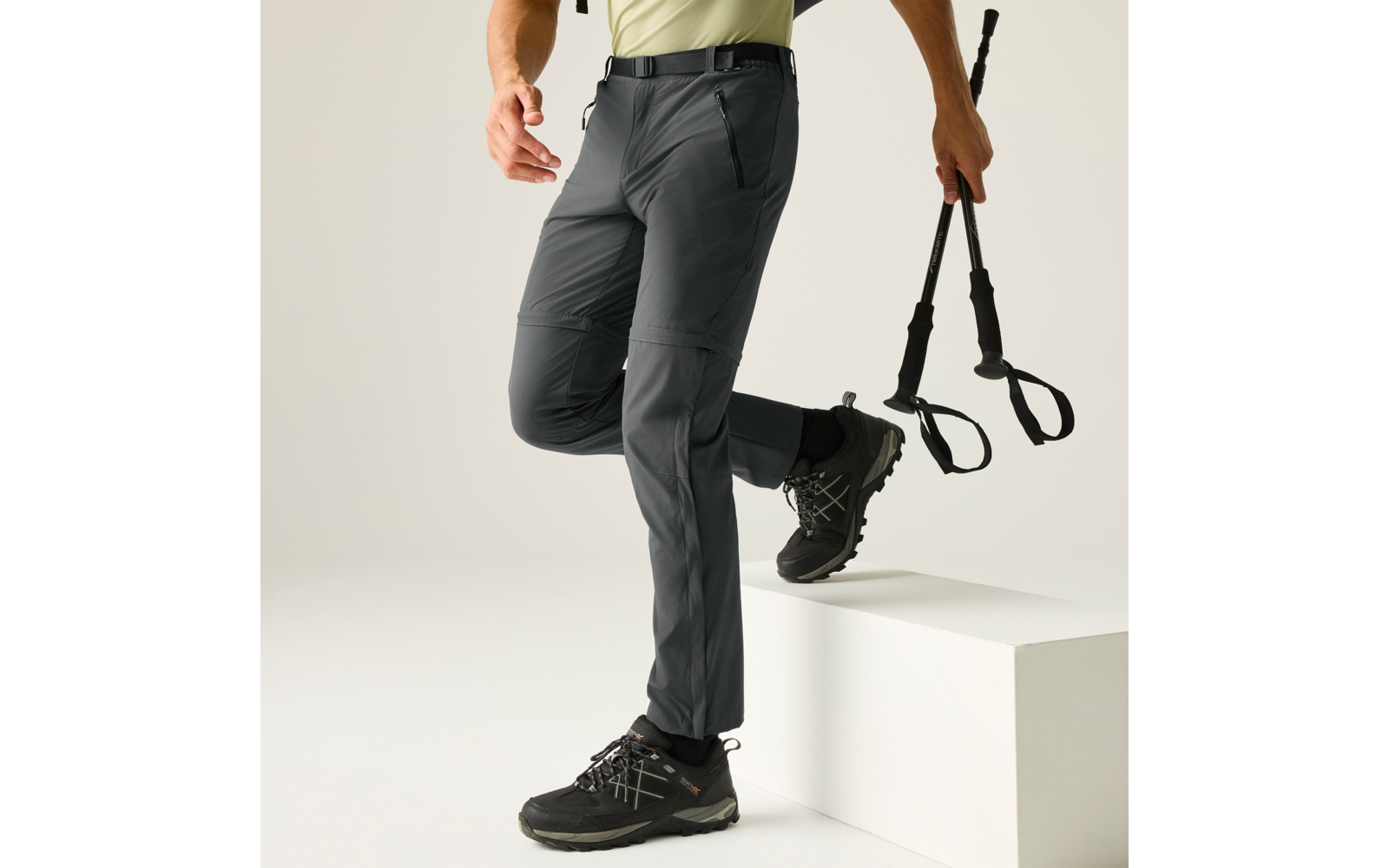 Unterkörper einer Person in grauer langen Hose und schwarzen, knöchelhohen Stiefeln; eine Hand hält zwei schwarze Stöcke mit Handschlaufen; ein Fuß steht auf einem weißen Block