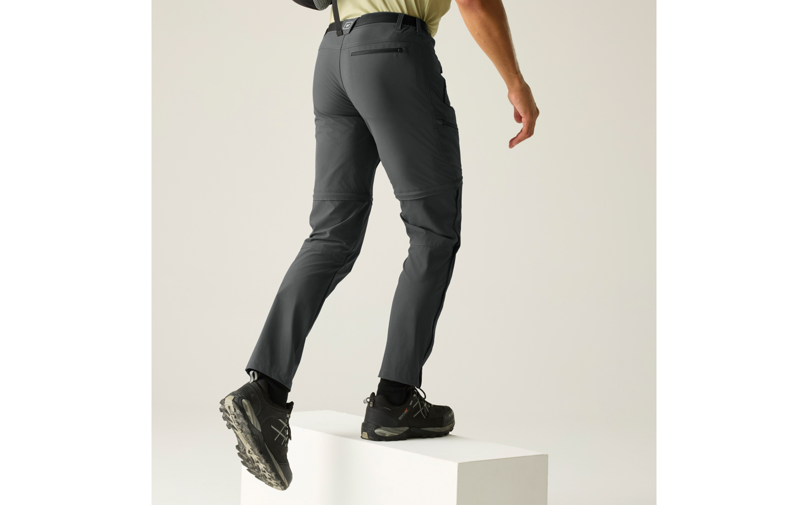 Unterkörper einer Person von hinten, hellgelbes Oberteil, dunkelgraue lange Hose mit Knienaht, Reißverschlusstasche am Gesäß und Oberschenkeltasche; eine Hand seitlich sichtbar; ein Fuß steht auf einem weißen Quader, der andere ist leicht angehoben; 