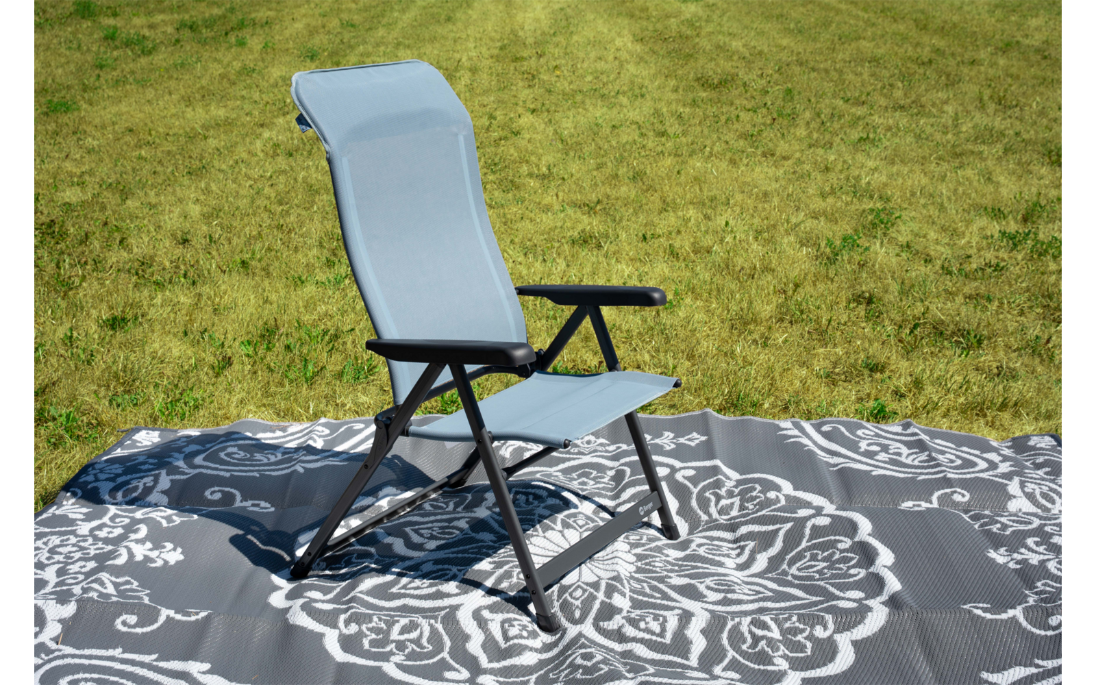 Sedia pieghevole azzurro chiaro con struttura nera e braccioli su un tappeto grigio con motivo floreale bianco, davanti a erba verde