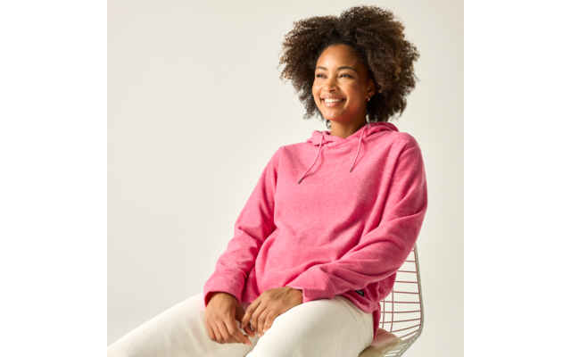Person mit lockigen Haaren sitzt auf einem Drahtstuhl, trägt pinken Hoodie und weiße Hosen, lächelt vor hellem Hintergrund