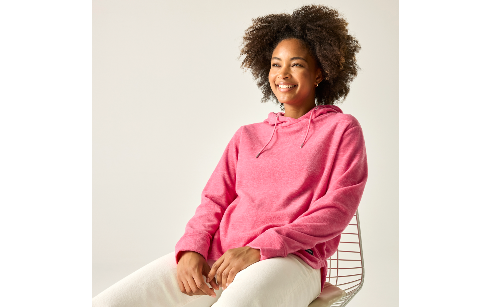 Person mit lockigen Haaren sitzt auf einem Drahtstuhl, trägt pinken Hoodie und weiße Hosen, lächelt vor hellem Hintergrund