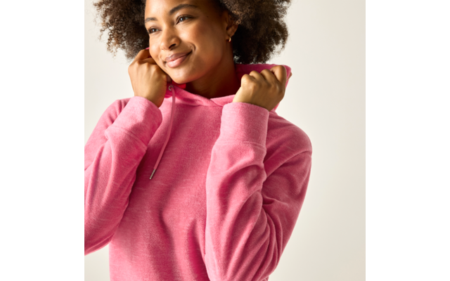 Person mit lockigem Haar trägt einen rosa Kapuzenpullover, hält die Kapuze am Kragen, schaut leicht nach rechts, leichtes Lächeln