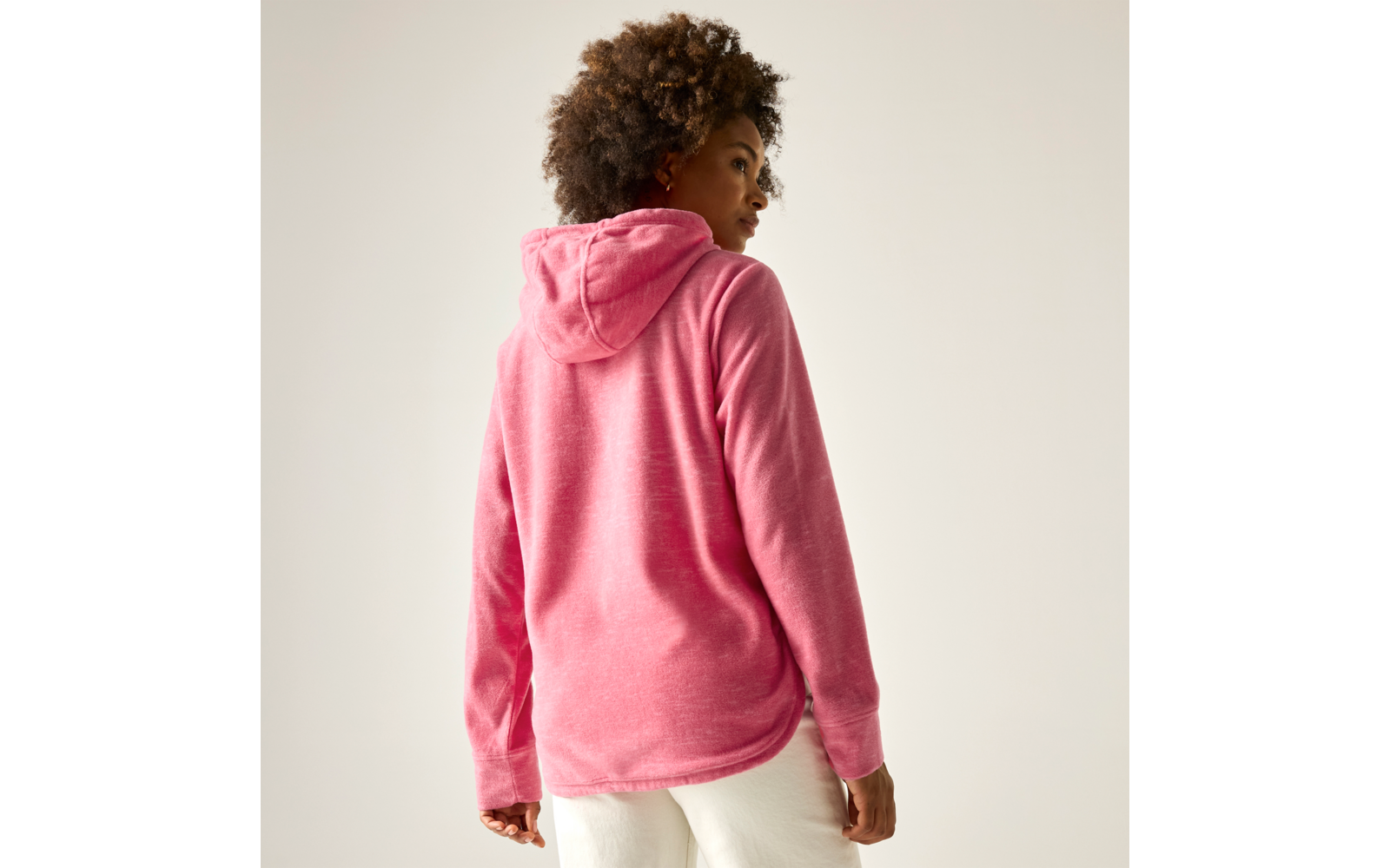 Person von hinten in rosa Kapuzenpullover und weißen Hosen, schaut über die rechte Schulter vor hellem, neutralem Hintergrund