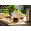 Vaso medidor de vidrio con marcas de medida, posos de café oscuros en el fondo; agua vertiéndose desde una tetera en el vaso; superficie de madera, planta en maceta, recipiente metálico y taza de cerámica en el fondo