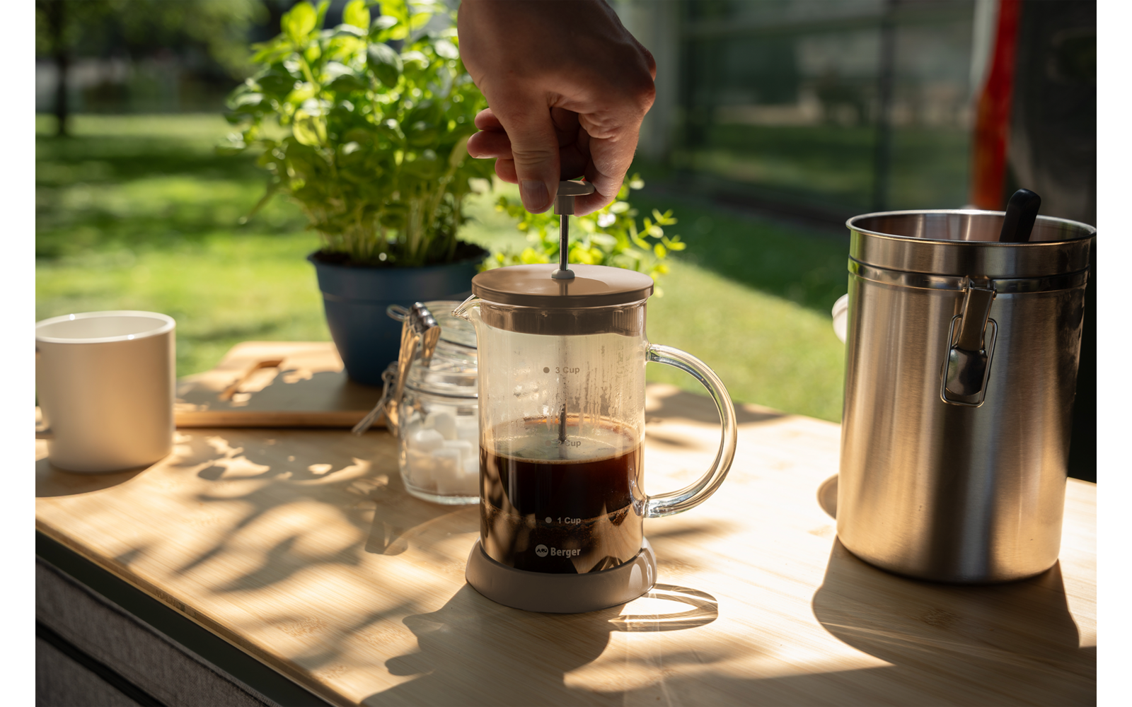 Mano presionando el émbolo metálico de una prensa francesa de vidrio con líquido oscuro sobre una superficie de madera; junto a ella una taza blanca, un vaso pequeño con contenido blanco, una jarra de leche de acero inoxidable y una planta en maceta, fondo de jardín desenfocado
