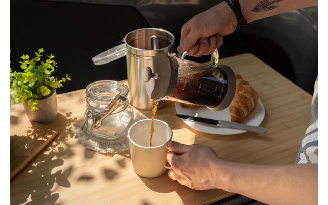 Mano vierte café de una prensa francesa en una taza sobre una mesa de madera; junto a ella un tarro de vidrio con tapa, una pequeña planta en maceta y un croissant en un plato con un cuchillo