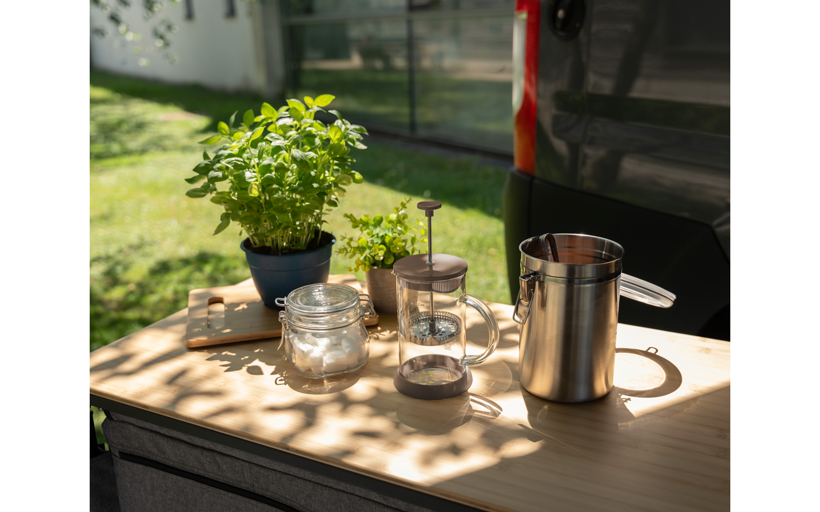 Mesa de madera al aire libre con dos macetas pequeñas con plantas verdes, dos tarros de vidrio, una cafetera de émbolo de vidrio y una jarra alta de acero inoxidable; lateral de un vehículo y césped al fondo