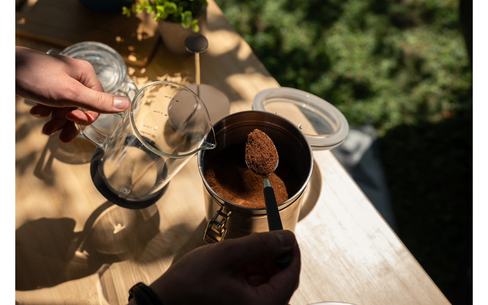 Dos manos: una sostiene una cuchara con polvo marrón sobre un recipiente metálico abierto, la otra sostiene un recipiente de vidrio con filtro metálico sobre una mesa de madera clara; al fondo plantas