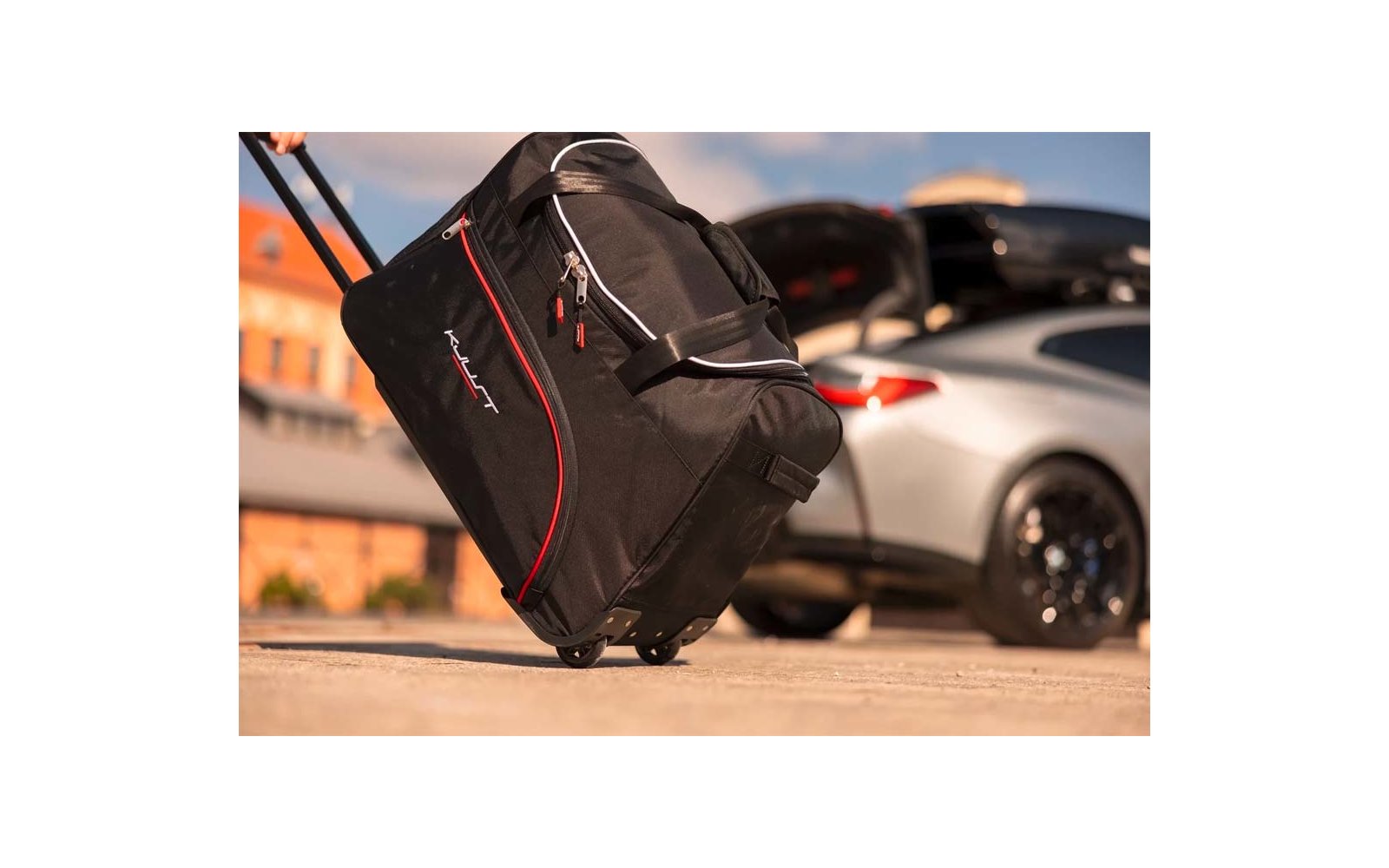 Sac de voyage noir à roulettes avec passepoil rouge et poignée télescopique sortie sur le bitume devant le coffre ouvert d'une voiture argentée