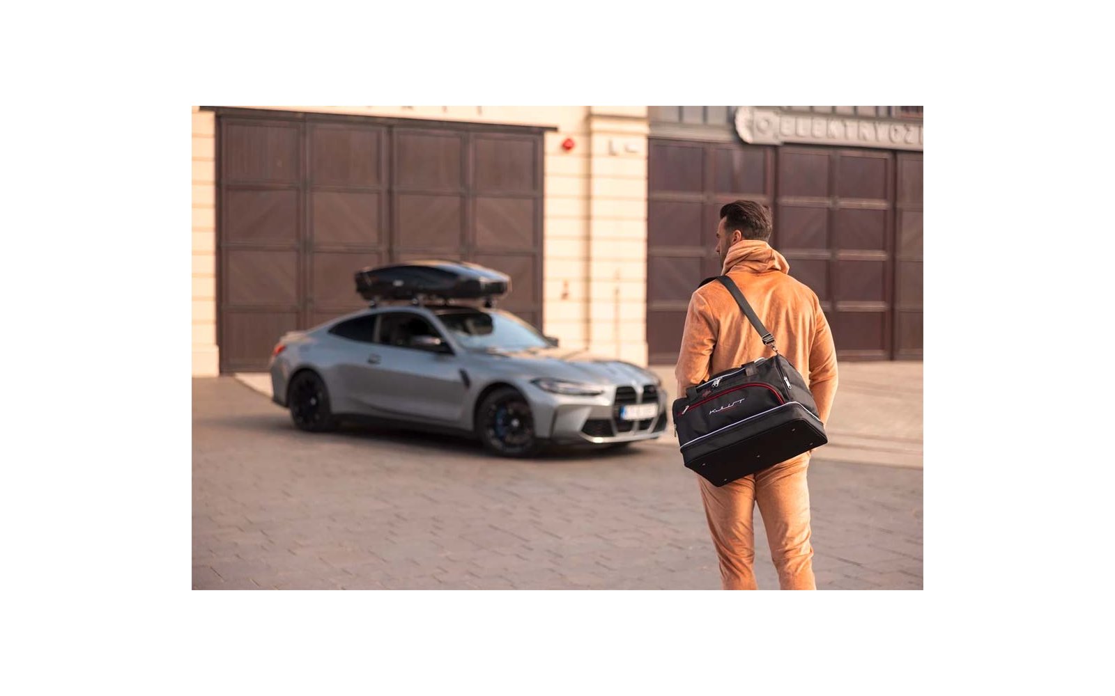 Homme vu de dos en sweat à capuche et pantalon beige, avec un sac noir à l'épaule, devant une voiture argentée garée avec coffre de toit; portes de garage brunes sur une façade claire en arrière-plan