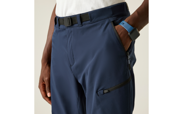 Plano detalle de pantalones azul oscuro con cinturón y bolsillo con cremallera en el muslo, borde de camiseta blanca visible; una mano en el bolsillo, la otra lleva un smartwatch con correa azul