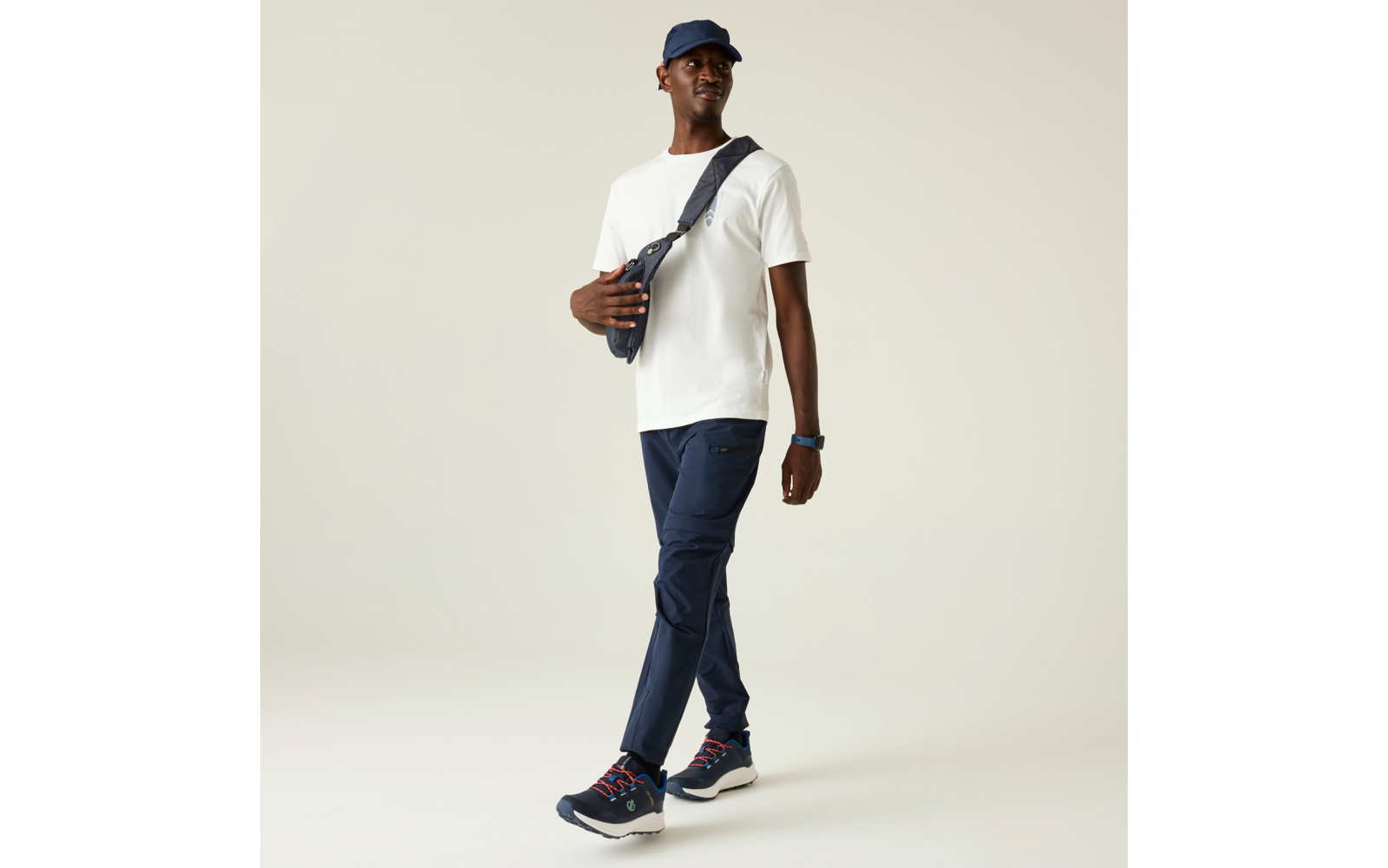 Persona caminando, viste camiseta blanca, pantalones azul oscuro, gorra oscura, bolso cruzado, zapatillas azul oscuro con suelas blancas, reloj de pulsera