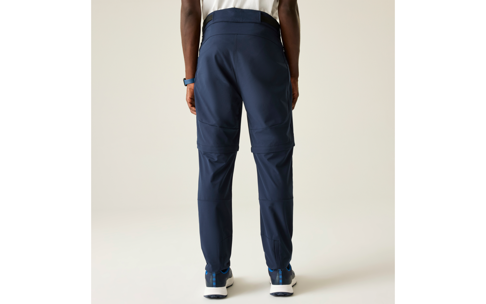 Vista posterior de una persona de pie de la cintura a los pies: pantalones azul oscuro con costuras horizontales a la altura de las rodillas, zapatillas blancas con detalles azules, camiseta blanca, reloj en la muñeca izquierda, fondo claro