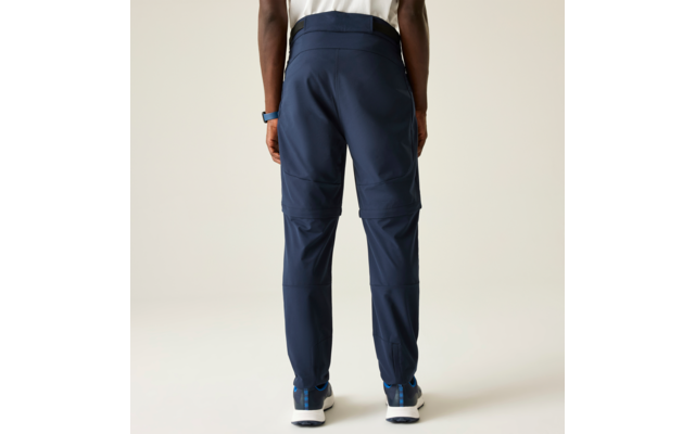 Vista posterior de una persona de pie de la cintura a los pies: pantalones azul oscuro con costuras horizontales a la altura de las rodillas, zapatillas blancas con detalles azules, camiseta blanca, reloj en la muñeca izquierda, fondo claro