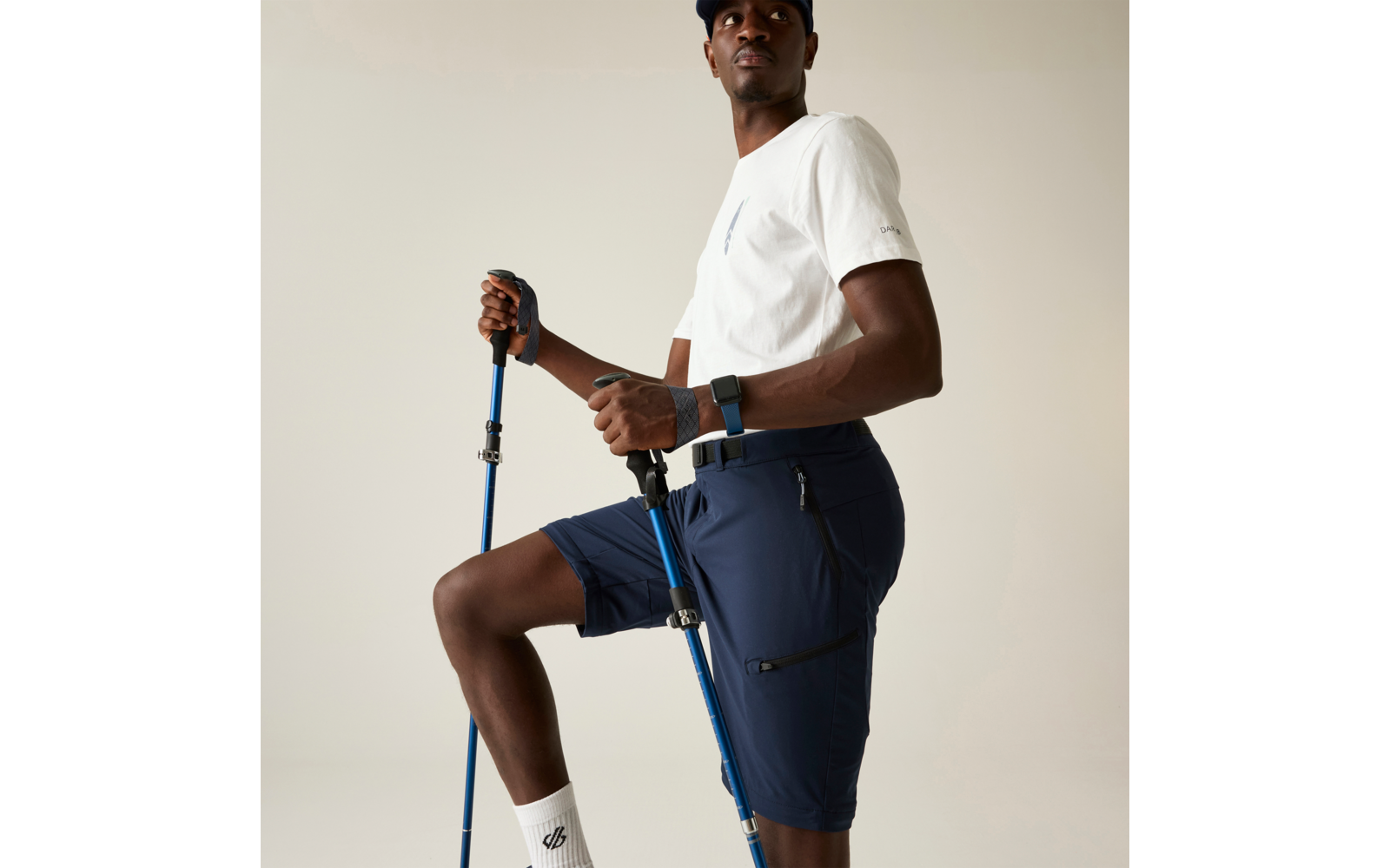 Persona con camiseta blanca y pantalones cortos azul marino sosteniendo dos bastones de senderismo azules, una rodilla levantada, fondo liso claro