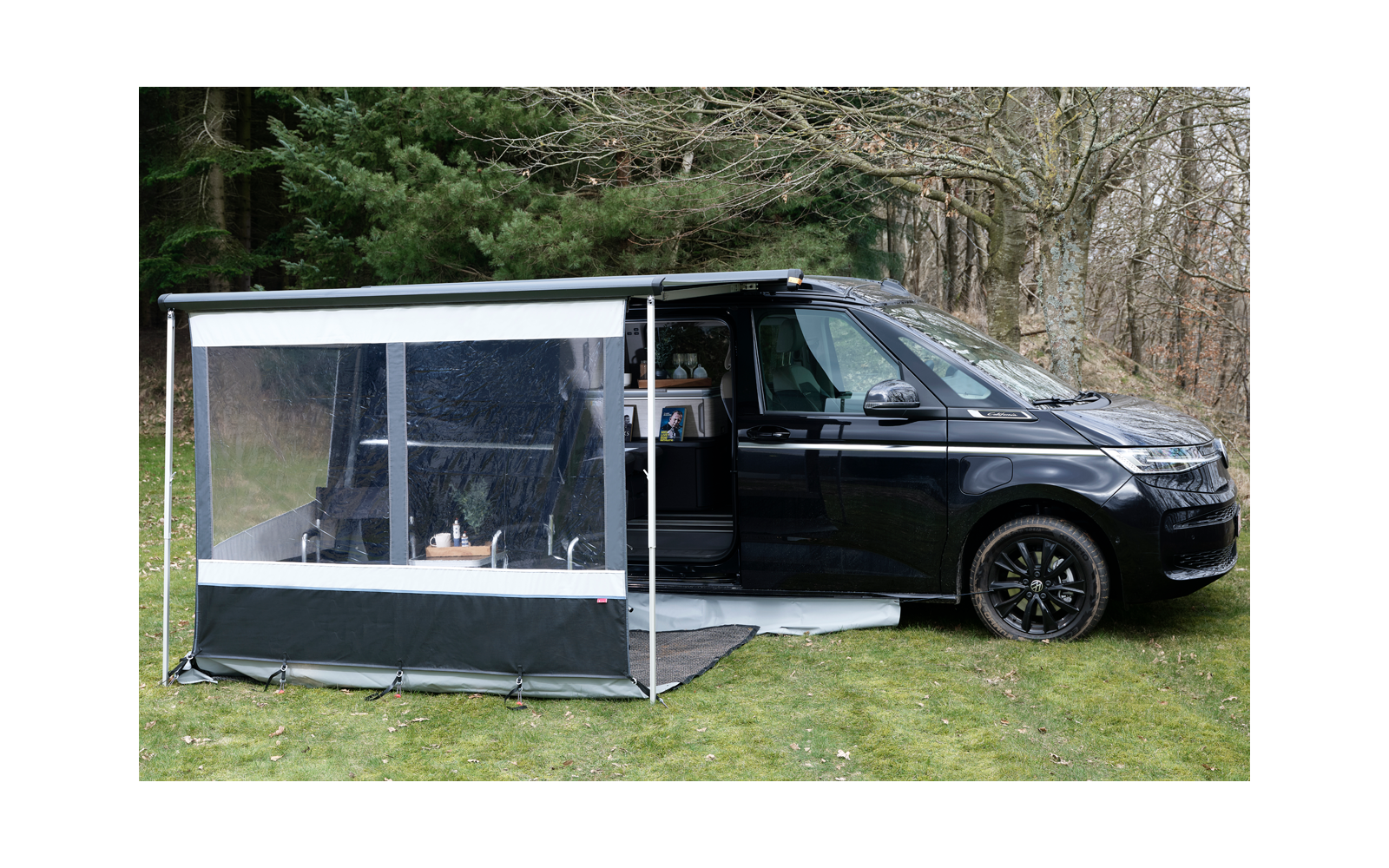 Schwarzer Campervan mit geöffneter Seitentür und angebautem transparenten Vorzelt auf einer Wiese vor Bäumen ohne Blätter