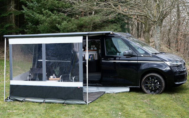 Schwarzer Campervan mit geöffneter Seitentür und angebautem transparenten Vorzelt auf einer Wiese vor Bäumen ohne Blätter