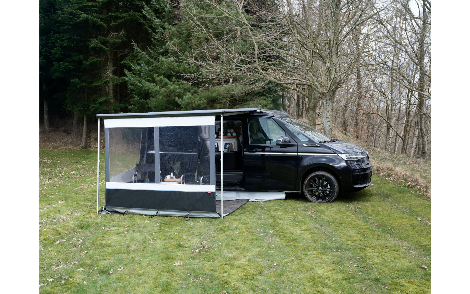 Schwarzer Campervan mit geöffneter Seitentür und angebautem transparenten Vorzelt auf einer Wiese vor Bäumen ohne Blätter