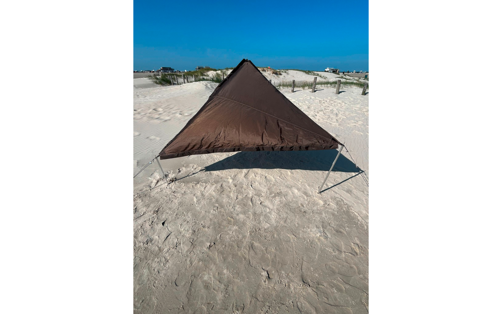 Dreieckiges braunes Sonnendach auf zwei Metallstangen auf sandigem Strand, Dünen mit Gras und blauer Himmel im Hintergrund
