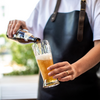 Person in weißem Shirt und schwarzer Schürze gießt Bier aus einer Flasche in ein Glas mit Schaum