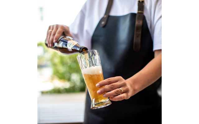Person in weißem Shirt und schwarzer Schürze gießt Bier aus einer Flasche in ein Glas mit Schaum