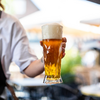 Hand hält ein hohes Bierglas mit goldenem Bier und dichter Schaumkrone, unscharfer Außenbereich im Hintergrund