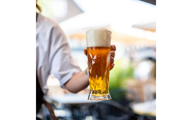 Hand hält ein hohes Bierglas mit goldenem Bier und dichter Schaumkrone, unscharfer Außenbereich im Hintergrund