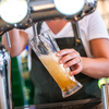 Person in weißem T‑Shirt und dunkler Schürze hält ein schräg gestelltes Bierglas unter einen Zapfhahn; Bier fließt, Schaum bildet sich