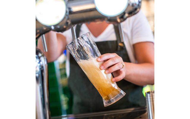 Person in weißem T‑Shirt und dunkler Schürze hält ein schräg gestelltes Bierglas unter einen Zapfhahn; Bier fließt, Schaum bildet sich