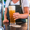 Hände einer Person in weißem T‑Shirt und dunkler Schürze halten ein hohes Bierglas unter einem Zapfhahn; Schaum läuft auf die Auffangfläche