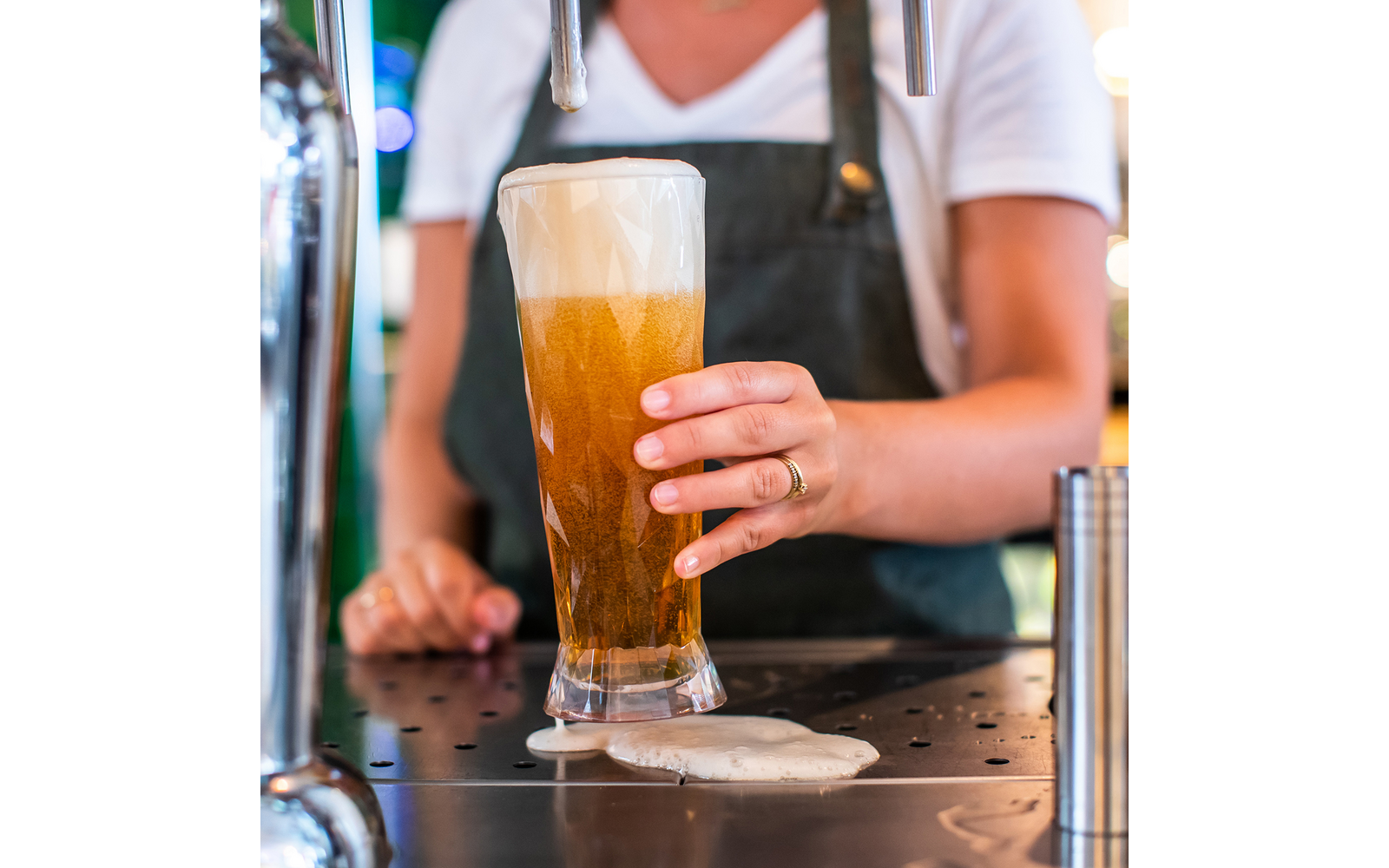 Hände einer Person in weißem T‑Shirt und dunkler Schürze halten ein hohes Bierglas unter einem Zapfhahn; Schaum läuft auf die Auffangfläche
