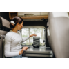 Donna con capelli raccolti in chignon e maglia bianca seduta in un camper, tocca un piccolo altoparlante nero su un tavolo; finestra con alberi sullo sfondo