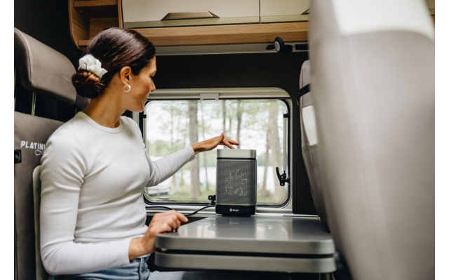 Donna con capelli raccolti in chignon e maglia bianca seduta in un camper, tocca un piccolo altoparlante nero su un tavolo; finestra con alberi sullo sfondo