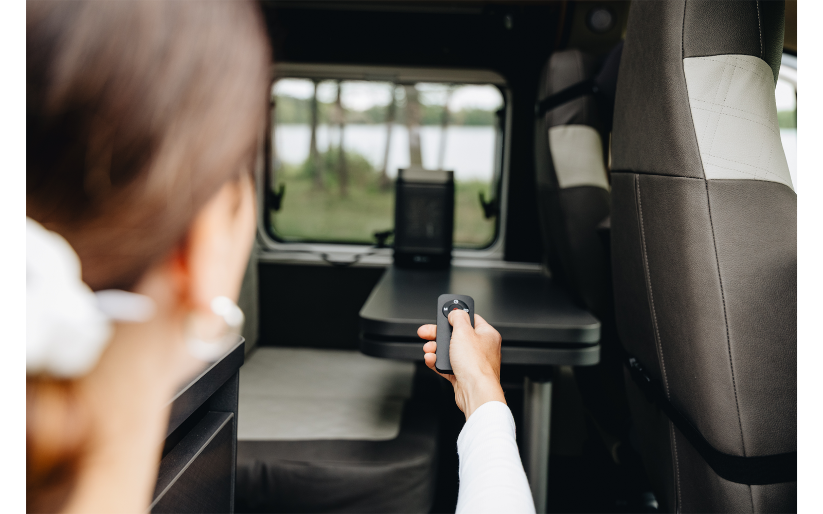 Persona vista da dietro nell'interno di un veicolo che tiene un telecomando puntato verso un tablet su un tavolo; finestra con alberi e acqua visibile