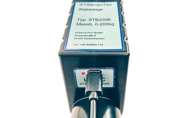 Nahaufnahme eines rechteckigen schwarzen Geräts mit weißem Etikett oben mit dem Text ATSensoTec Stabwaage Typ. STB200B Messb. 0-200kg und einem schwarzen Kabel, das in eine Öffnung an der Vorderseite gesteckt ist
