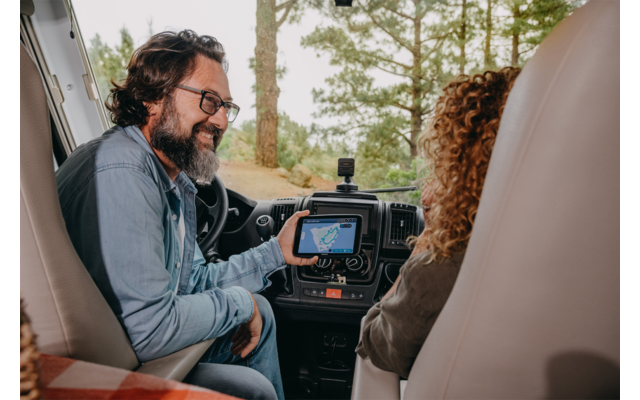 Mann mit Brille auf dem Fahrersitz eines Wohnmobils zeigt auf ein Navigationsgerät; Frau mit lockigen Haaren sitzt auf dem Beifahrersitz; Bäume sind durch die Windschutzscheibe sichtbar