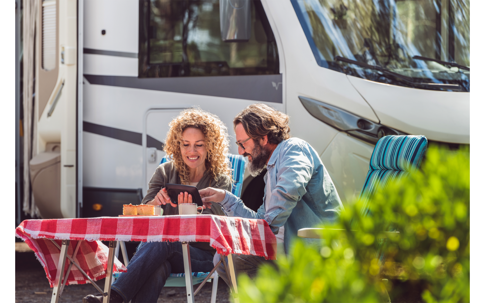 Zwei Erwachsene sitzen an einem kleinen Tisch mit rot-weiß kariertem Tischtuch vor einem weißen Wohnmobil und schauen auf ein Tablet; Tassen auf dem Tisch, eine Person trägt eine Sonnenbrille auf dem Kopf, im Vordergrund ein grüner Strauch