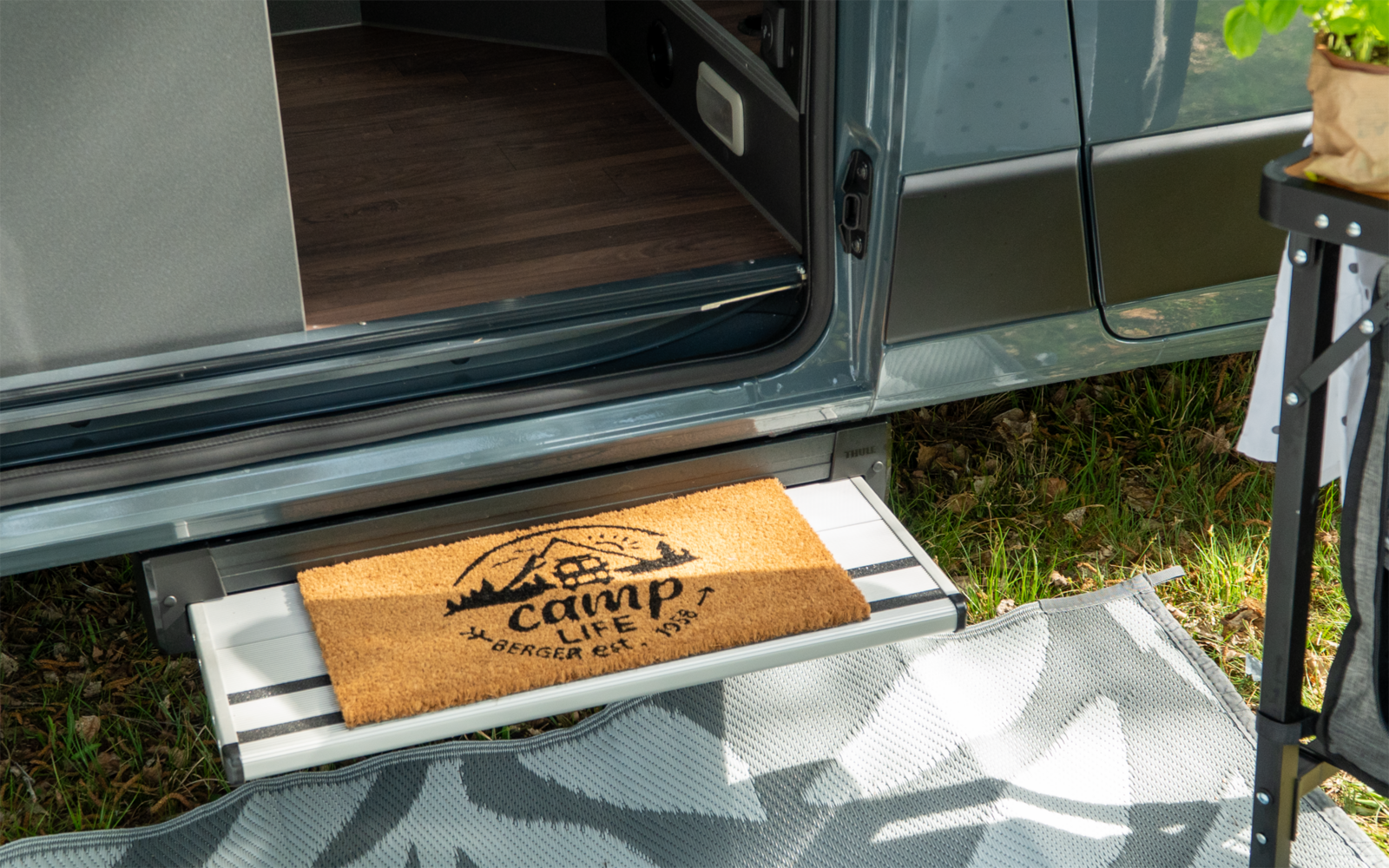 Porte de véhicule ouverte avec marche métallique ; sur la marche un paillasson en coco avec l'inscription 'camp life' et un motif de montagne ; devant un tapis d'extérieur à motif, de l'herbe et un pied de table