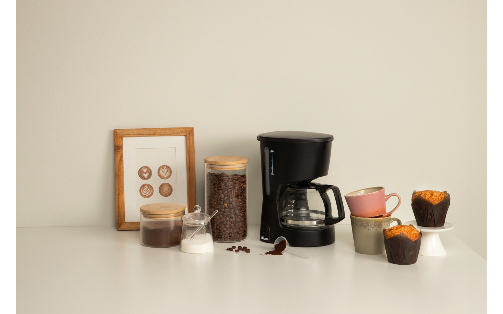 Machine à café filtre noire avec carafe en verre sur une surface blanche ; à gauche un bocal en verre contenant des grains bruns, petits récipients en verre avec contenu blanc et brun, grains éparpillés et une cuillère en bois ; à droite des tasses empilées et trois muffins sur de petits présentoirs ; en arrière‑plan un cadre appuyé contre le mur