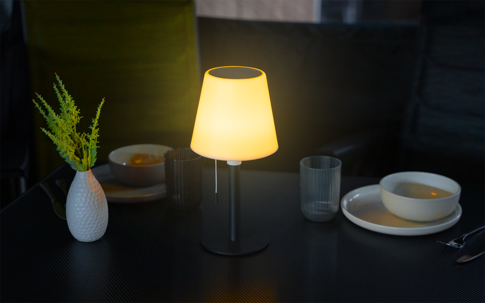 Beleuchtete Tischlampe auf dunklem Esstisch, daneben zwei Teller mit Schalen, zwei Gläser, Besteck und eine weiße Vase mit grünen Zweigen