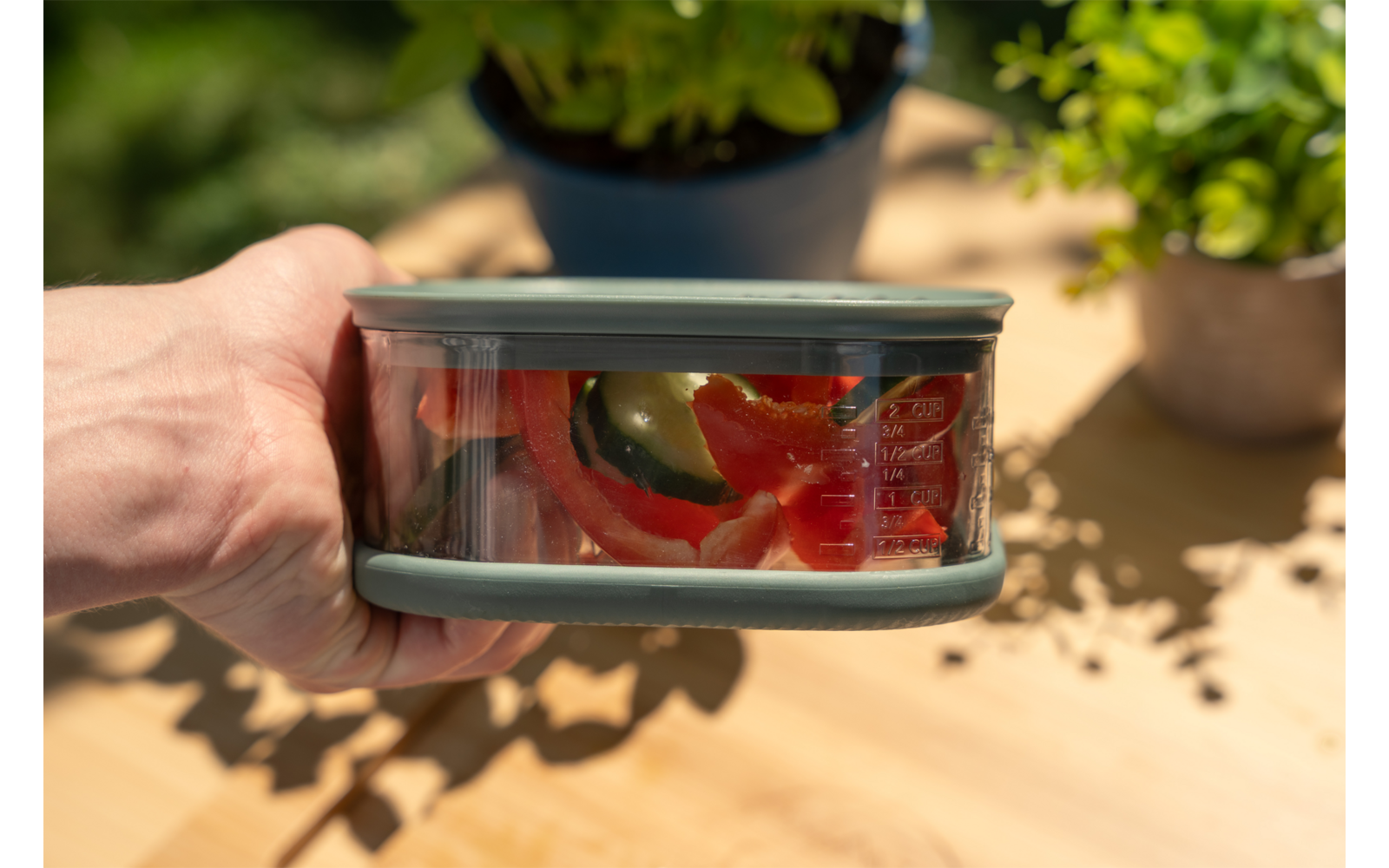 Hand houdt doorzichtige bewaardoos met grijs deksel met daarin gesneden rode paprika en groene groenteschijfjes; houten tafel en twee bloempotten op de achtergrond