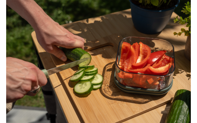 Twee handen gebruiken een mes om een komkommer op een houten snijplank te snijden; daarnaast een glazen bak met gesneden rode paprika, een hele komkommer en potplanten op de achtergrond