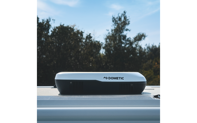 Weißes rechteckiges Gerät mit Schriftzug "DOMETIC" auf einer geriffelten hellgrauen Dachfläche, im Hintergrund Bäume und blauer Himmel