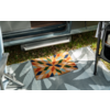 Tapis de sol rectangulaire coloré avec motif géométrique en forme d'étoile sur un tapis d'extérieur gris devant une porte de véhicule ouverte ; marche en métal, herbe et partie d'une structure métallique visibles