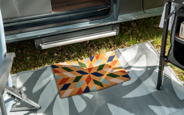 Tapis de sol rectangulaire coloré avec motif géométrique en forme d'étoile sur un tapis d'extérieur gris devant une porte de véhicule ouverte ; marche en métal, herbe et partie d'une structure métallique visibles